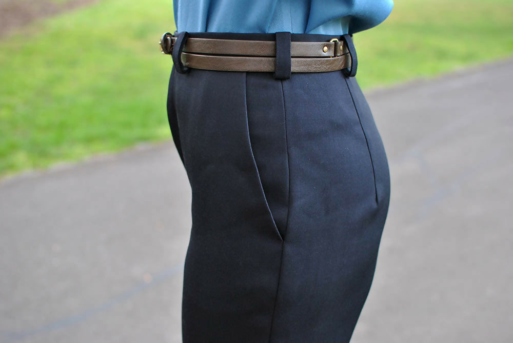 Details of a black formal pants. A woman is wearing them with blue blouse and brown belt.