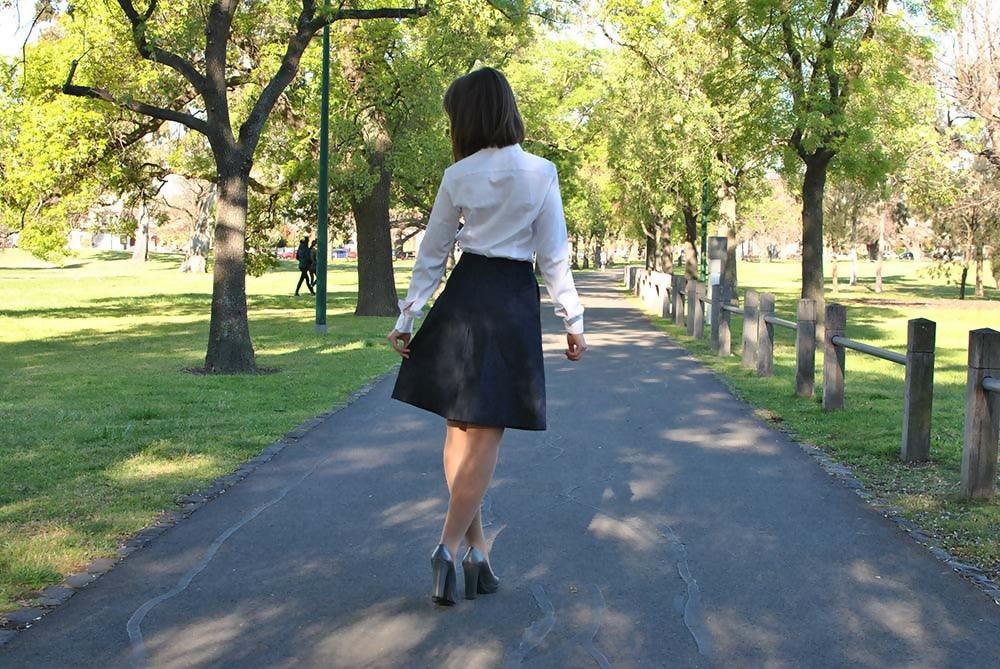 The back side of a woman who is wearing navy bue denim coulotte skirt, white shirt and gray high heel shoes while she is standing on a road in a park.