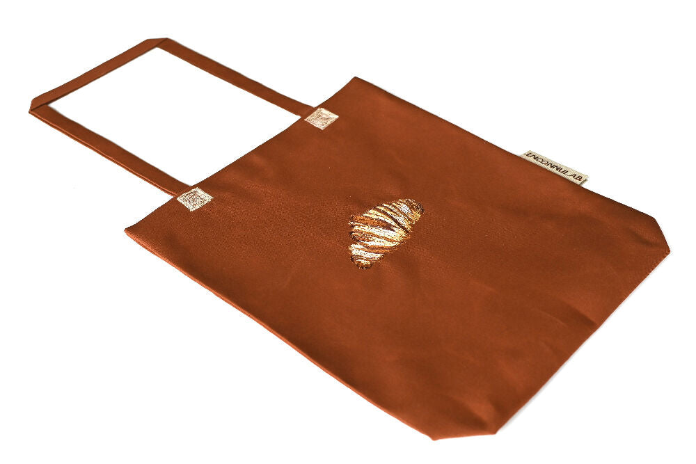brown paperbag lookalike canvas tote with croissant embroidery on it lying on a white table.
