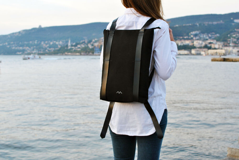 woman in white shirt is standing in front of the sea while wearing a full black minimalist backpack.