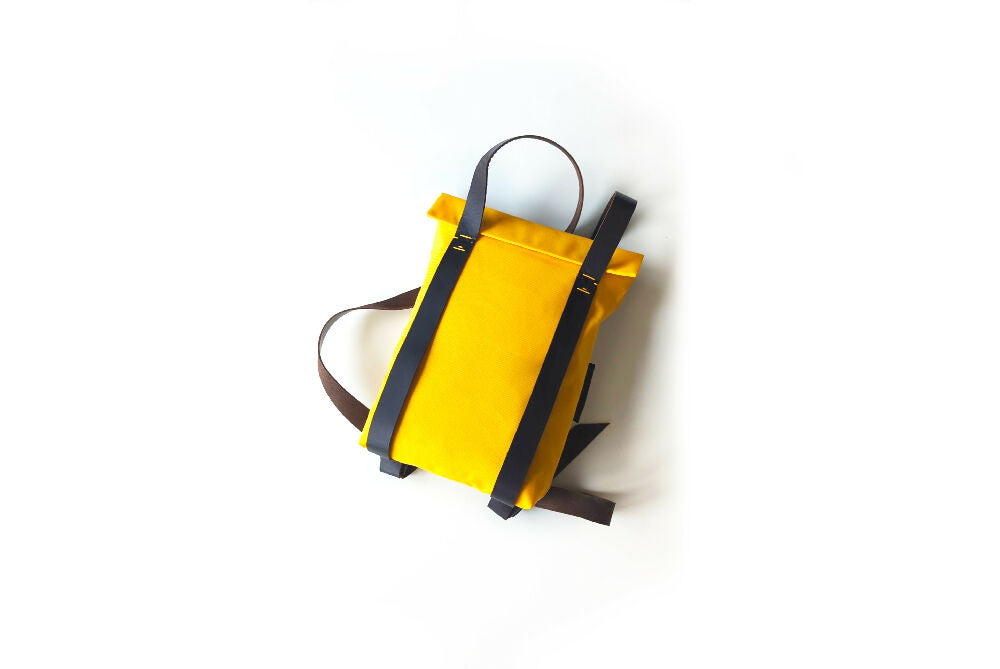 A bright yellow canvas backpack with black leather straps is lying on a white surface.