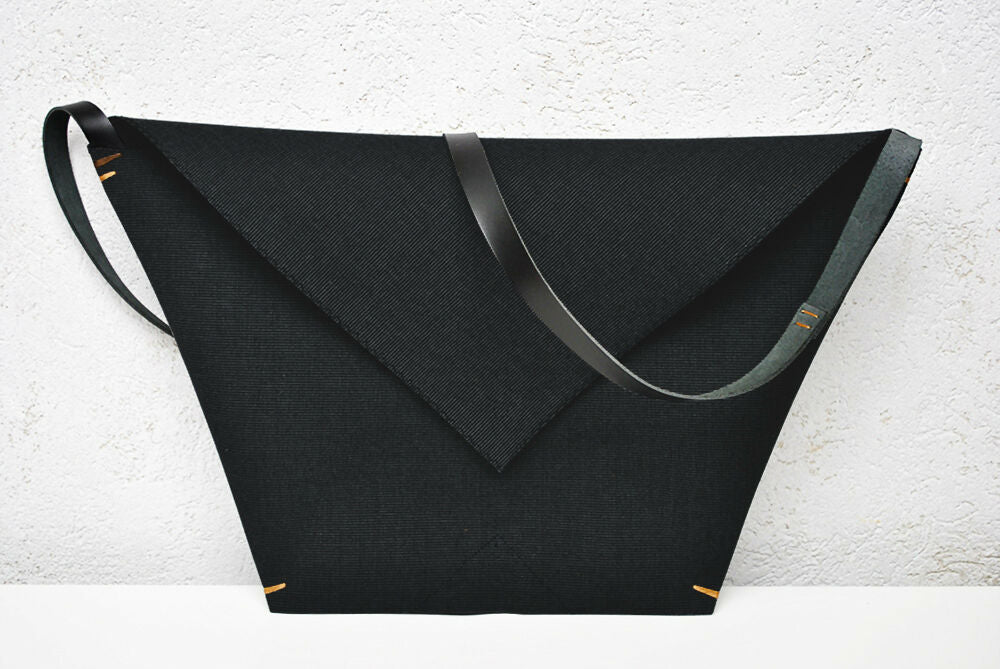 Black minimalist envelope bag with black leather strap is standing on a white table.