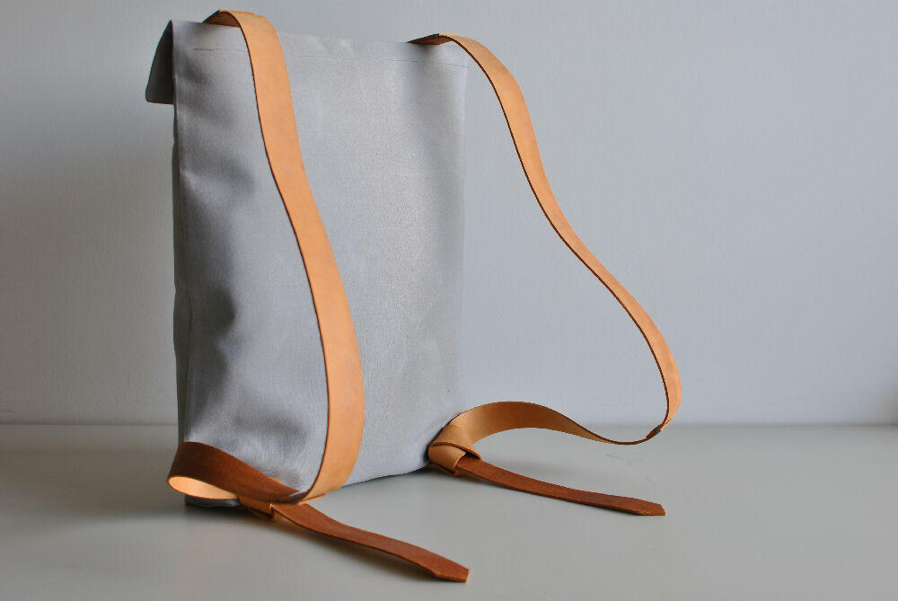 the backside of a grey minimalist backpack with nude leather straps is standing on a beige table in front of a white wall. The straps finish in a knot.