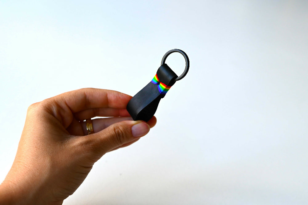 A hand is holding a black leather keychain with colourful rainbow stitches and black split ring. White background.