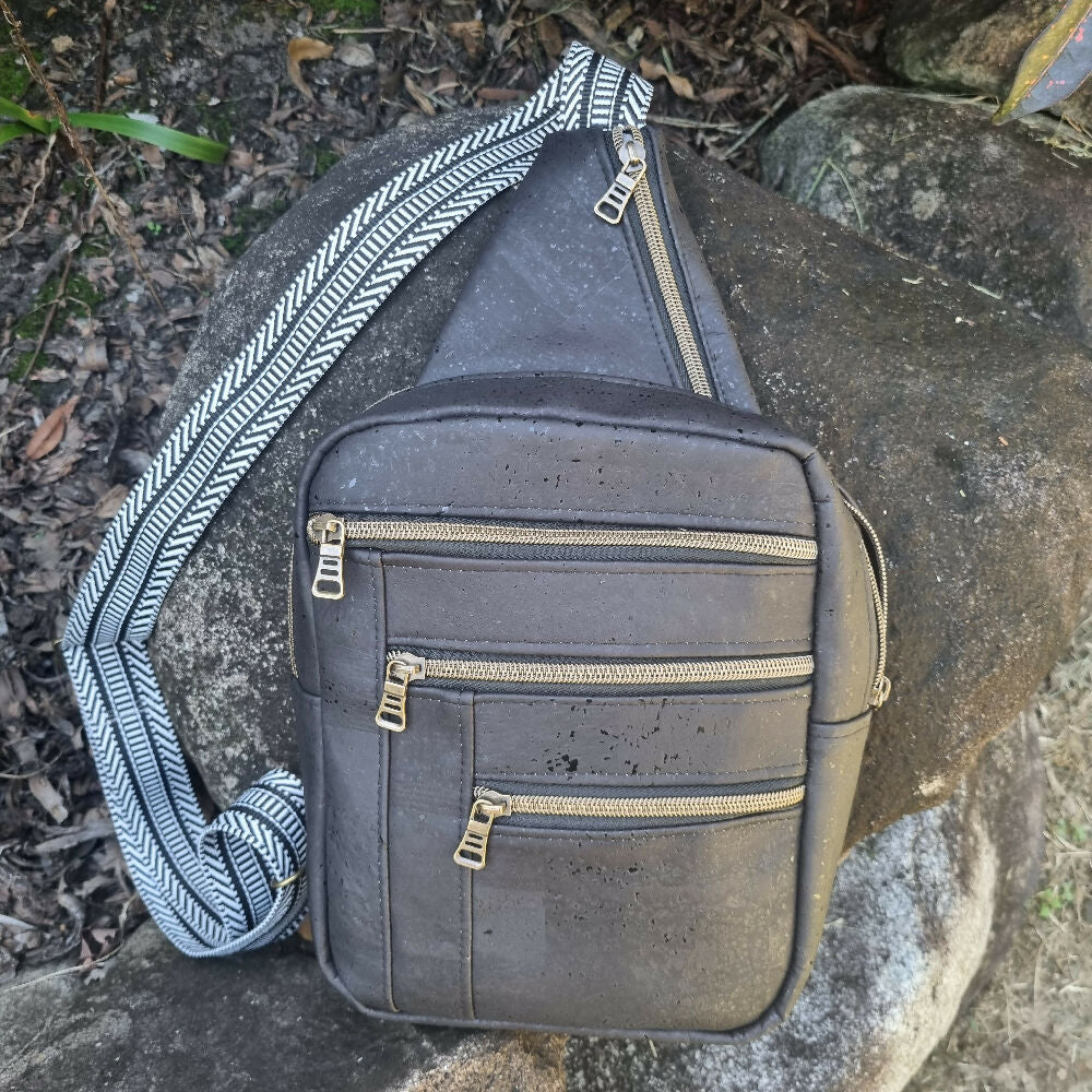 Black cork sling bag, front view showing four zipped pockets and decorative webbing.