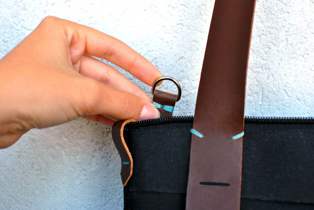A hand is holding a brown leather keychain of a black canvas bag with zipper closure.