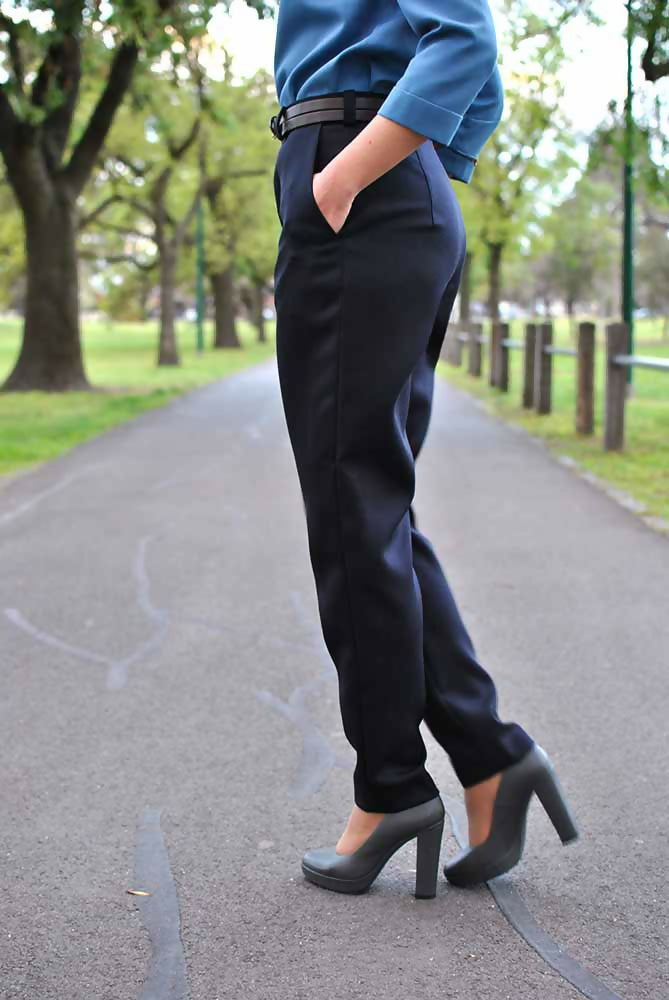 The lower part of a woman who is wearing black formal pants, blue blouse and gray high heel shoes. Her hands are in her pants' pockets.