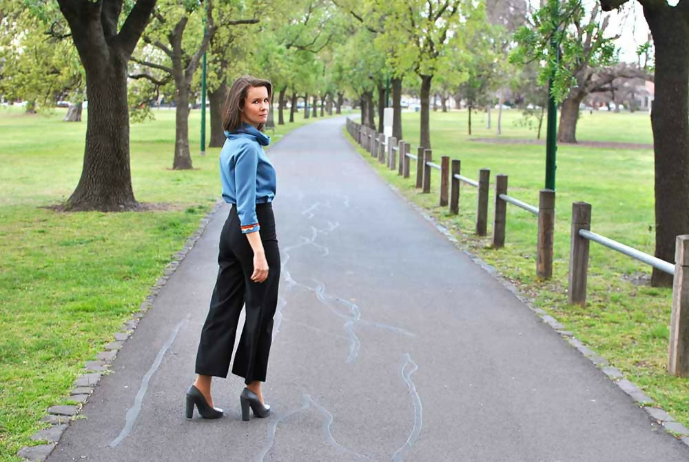 Woman in black wool crop pants and blue blouse is looking back on a road in a park.