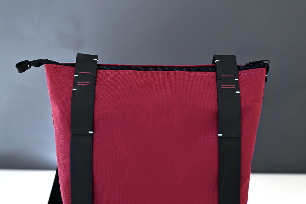 the upper side of a bordeaux colour canvas zipper backpack with black handstitched leather straps.