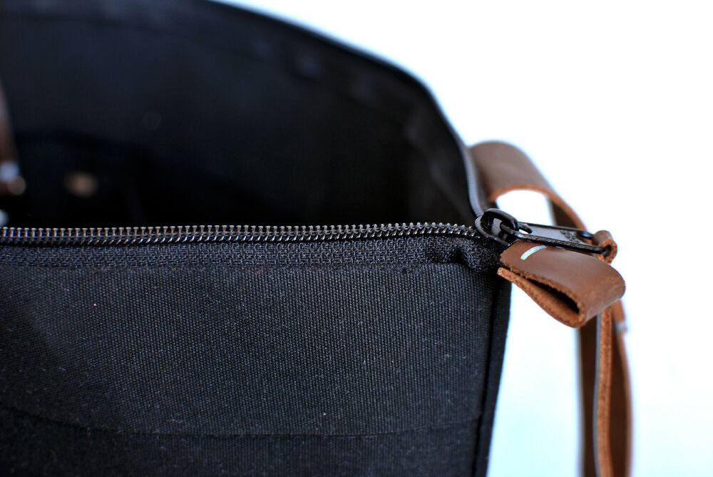 close-up photo of a black canvas bag with zipper cosure and brown leather strap. The inside of the bag is visible too.