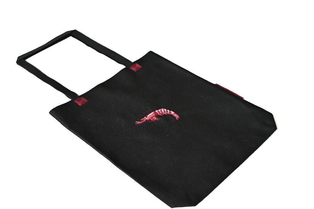 Black paperbag lookalike canvas tote with prawn embroidery on it lying on a white table.