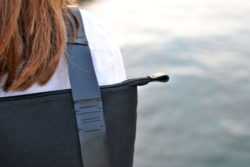 close-upe photo of the shoulder of a women who is wearing white shirt and black canvas backpack with black handstitched leather straps. water in the background.