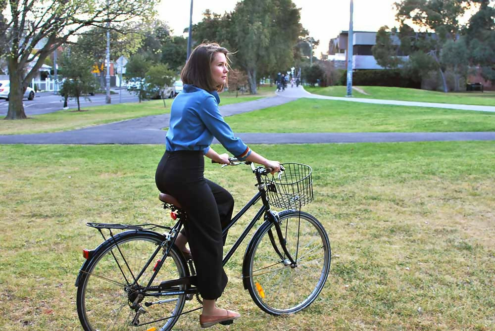 Woman who is wearing black palazzo pants, blue blouse and brown shoesis cycling with a black Dutch style bicycle.