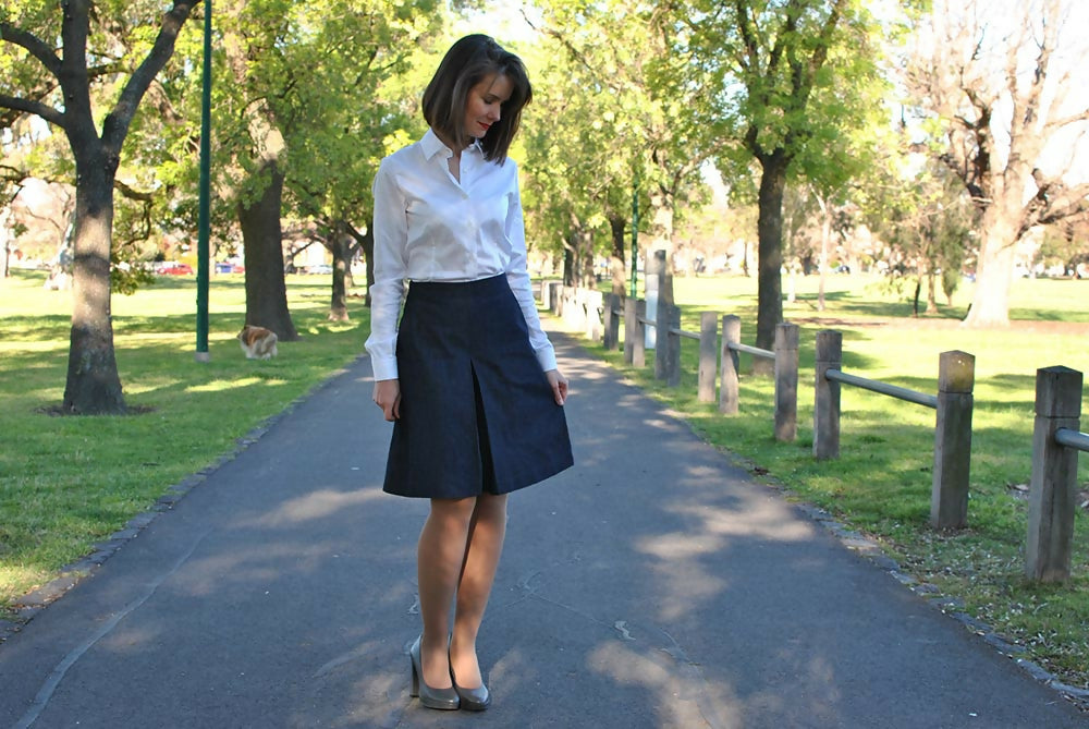 A woman is wearing navy bue denim coulotte skirt, white shirt and gray high heel shoes while she is standing on a road in a park.