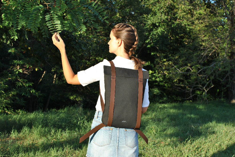 woman in white shirt and denim skirt with black minimalist backpack and brown leather straps is reaching out to grasp a little twig.