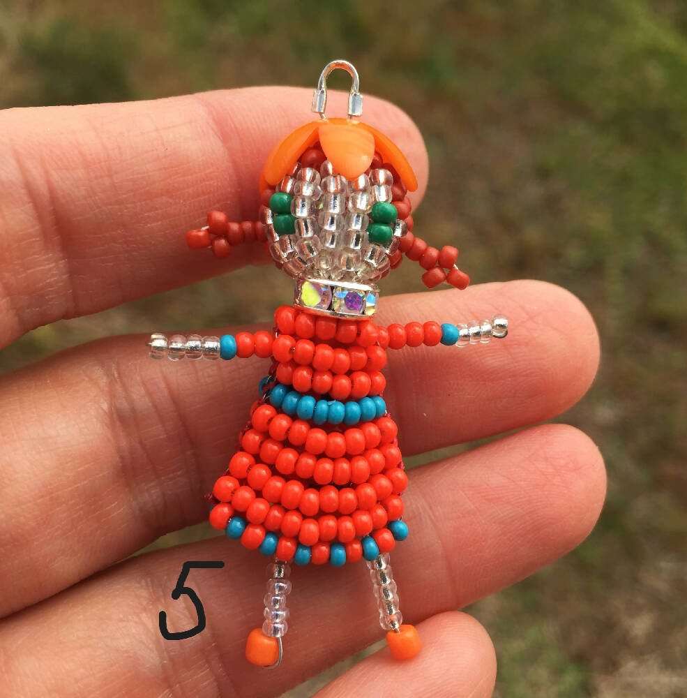 Naryanabeads beaded doll option 5. Beaded doll with clear crystal collar, orange flower bead hat, light brown braided hair and emerald eyes. Legs, arms, face made of shiny clear beads, orange-light blue dress. silver colour loop on top of hat