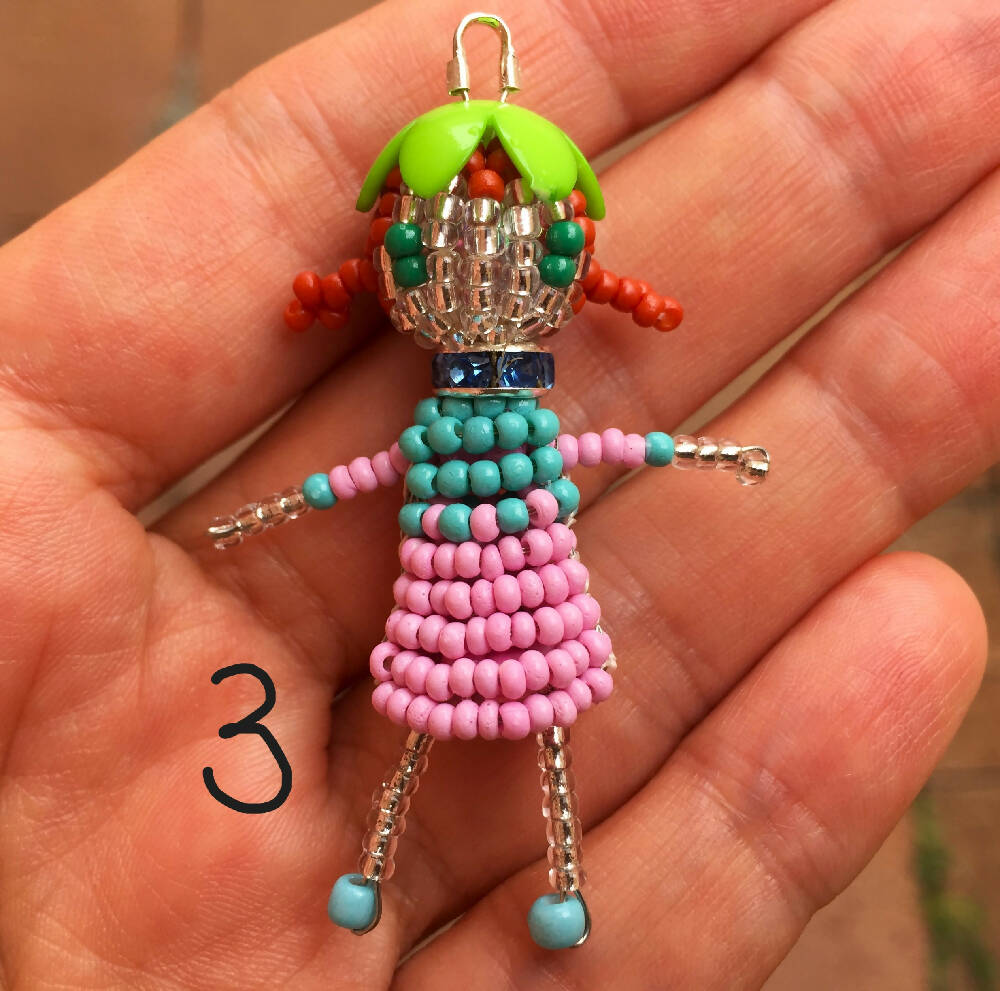 Naryanabeads beaded doll option 3d doll. Beaded doll with blue crystal collar, green flower bead hat, light brown braided hair and emerald eyes. Legs, arms, face made of shiny clear beads, pink-light blue dress. silver colour loop on top of hat