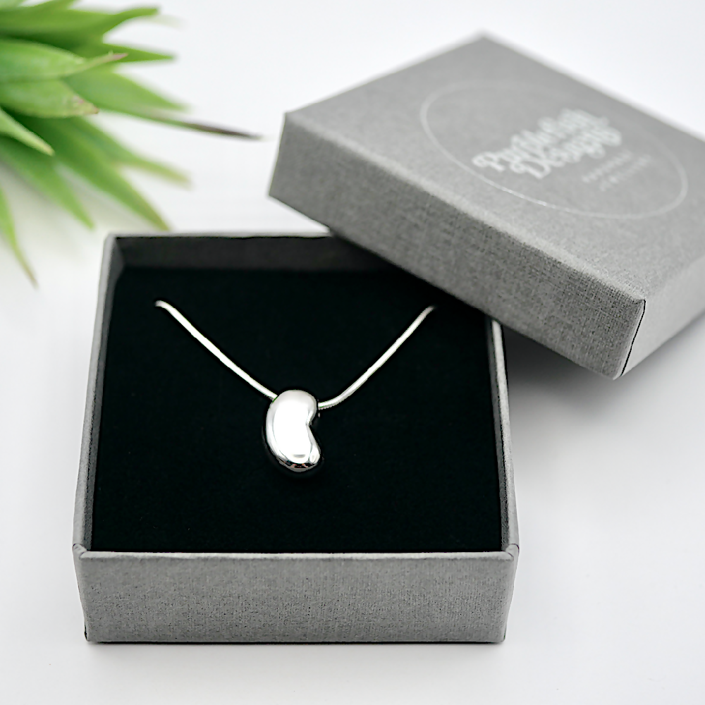 Image of handmade sterling silver bean shaped pendant on a snake chain in a linen textured grey jewellery gift box embossed with silver Purplefish Designs Handmade Jewellery logo. Box is on a white background with a decorative green plant.