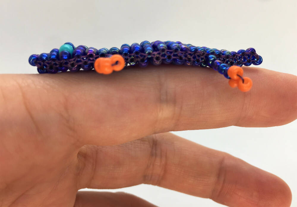 On the pointing finger shiny dark blue beaded crocodile showing the side of the body. Body made of shine dark blue seed beads, bright blue seed bead eyes, orange seed bead paws