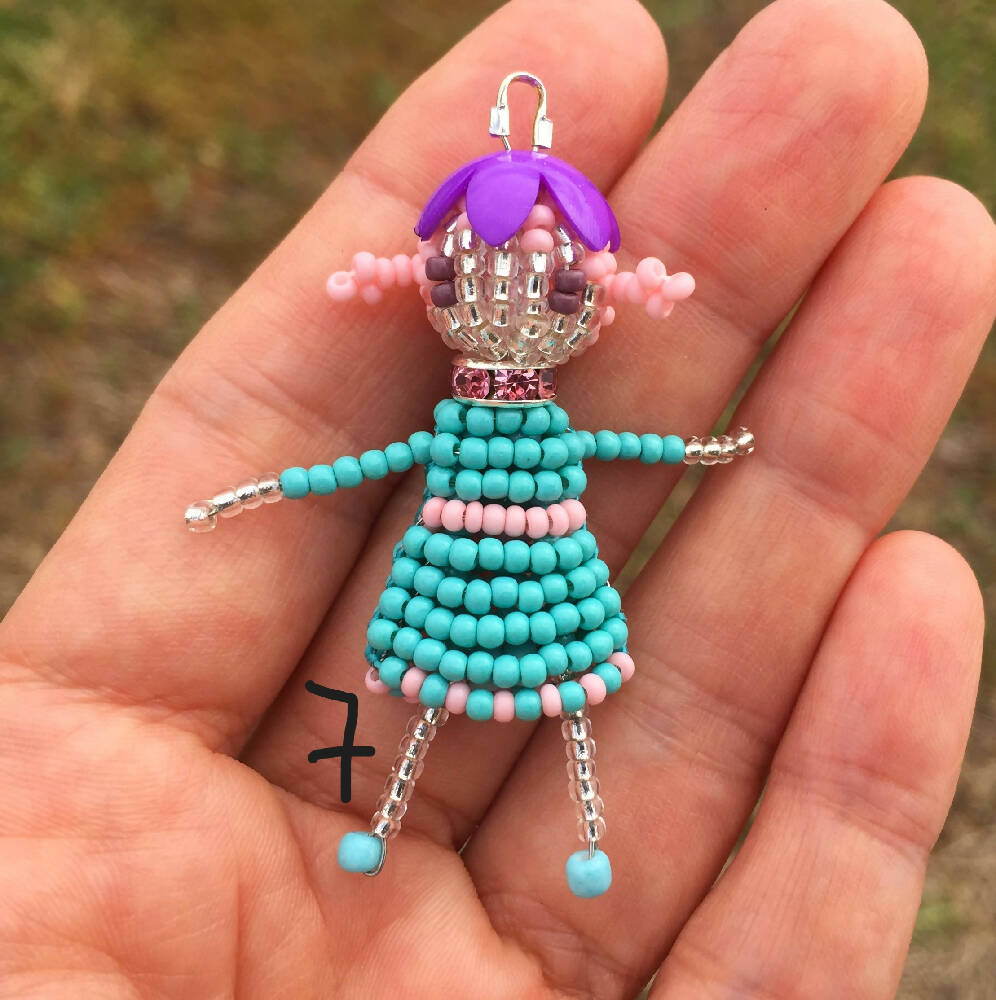 Naryanabeads beaded doll option 7. Beaded doll with  crystal collar, purple flower bead hat, pink braided hair and brown eyes. Legs, arms, face made of shiny clear beads, light blue-pink dress. silver colour loop on top of hat