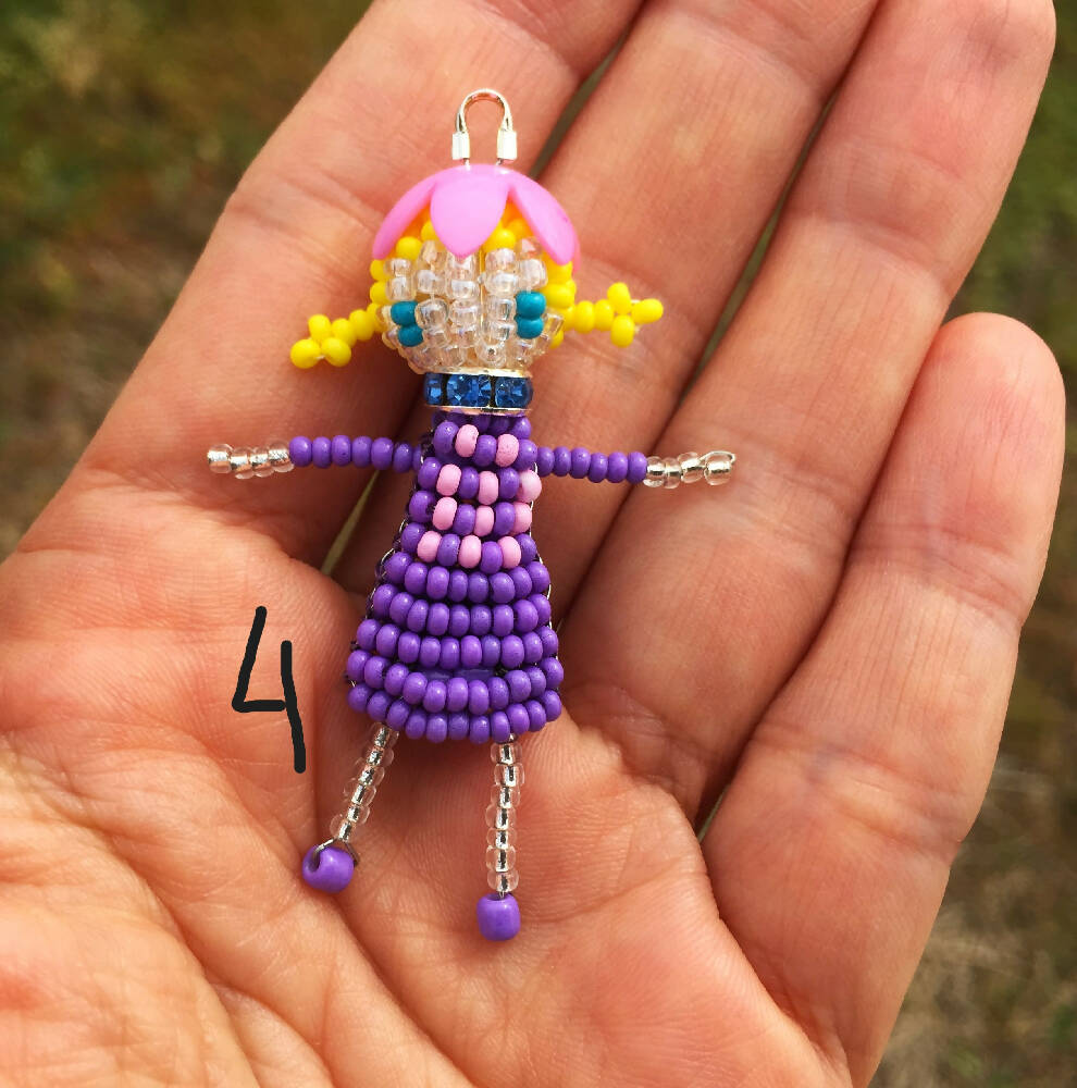 Naryanabeads beaded doll option 4. Beaded doll with blue crystal collar, pink flower bead hat, yellow braided hair and light blue eyes. Legs, arms, face made of shiny clear beads, purple-pink dress. silver colour loop on top of hat