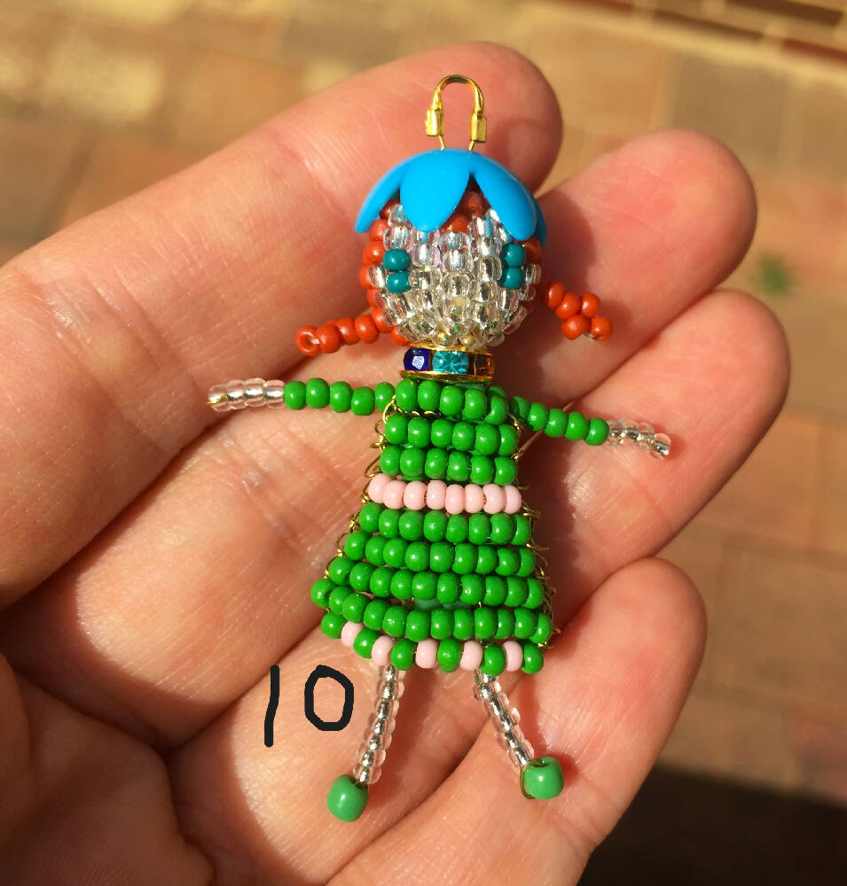 Naryanabeads beaded doll option 10. beaded doll with multicolour  crystal collar, green flower bead hat, light brown braided hair and emerald eyes. Legs, arms, face made of shiny clear beads, green-pink dress. golden colour loop on top of hat