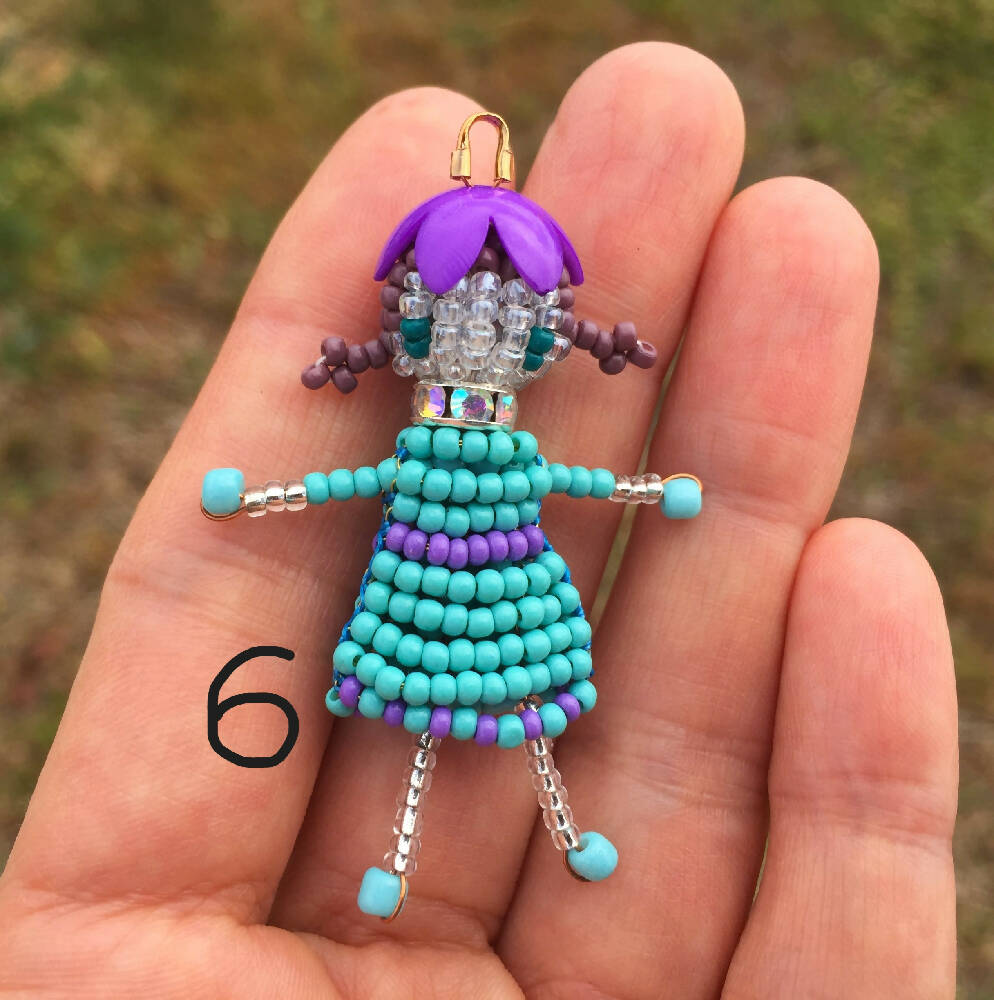 Naryanabeads beaded doll option 6. Beaded doll with clear crystal collar, purple flower bead hat, light brown braided hair and emerald eyes. Legs, arms, face made of shiny clear beads, blue dress with purple trim. silver colour loop on top of hat
