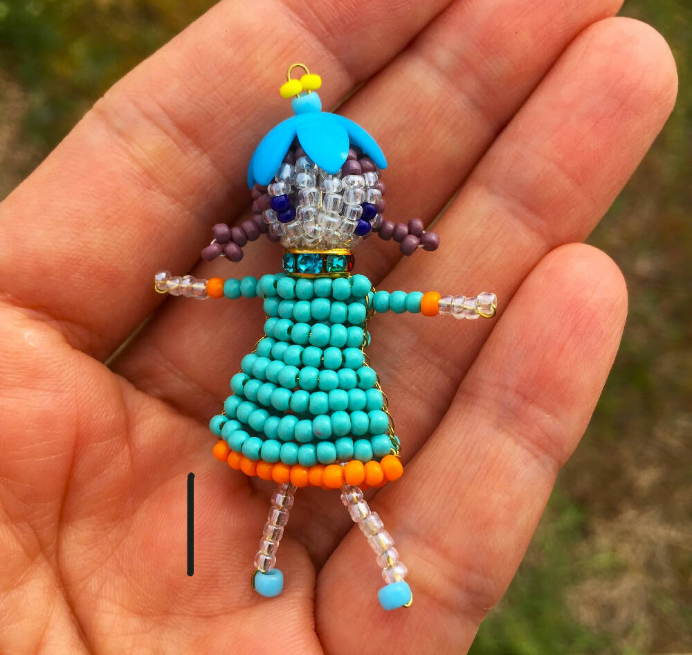 Naryanabeads beaded doll option1 - beaded doll with light blue crystal collar, blue flower bead hat, brown braided hair and ink blue eyes. Legs, arms, face made of shiny clear beads, light blue dress with orange trim.