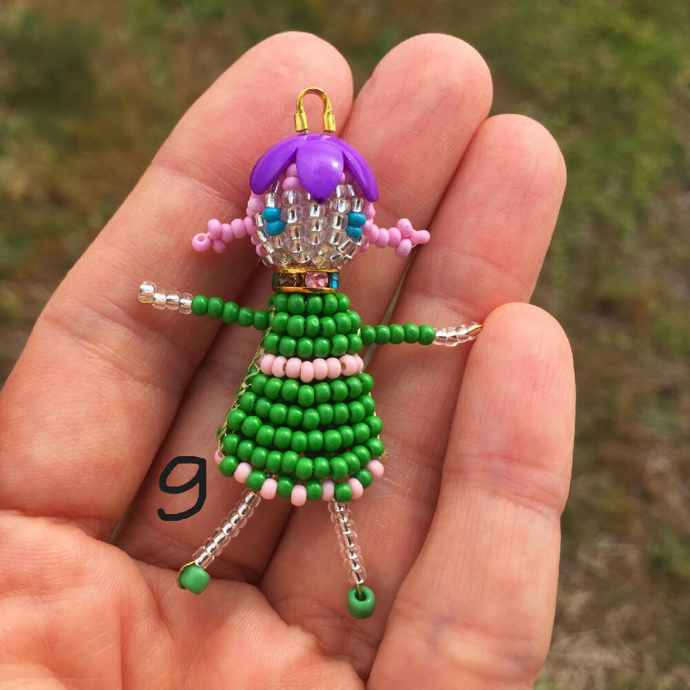 Naryanabeads beaded doll option 9. Beaded doll with multicolour  crystal collar, purple flower bead hat, pink braided hair and blue seed eyes. Legs, arms, face made of shiny clear beads, green dress with pink trim. golden colour loop on top of hat