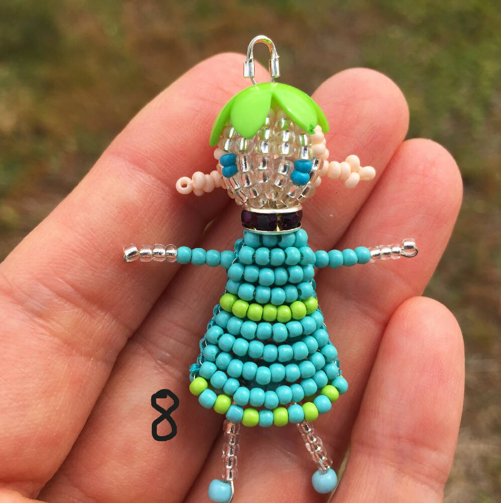 Naryanabeads beaded doll Option 8. Beaded doll with crystal collar, green flower bead hat, light peach braided hair and ligh blue eyes. Legs, arms, face made of shiny clear beads, light blue-green dress. silver colour loop on top of hat