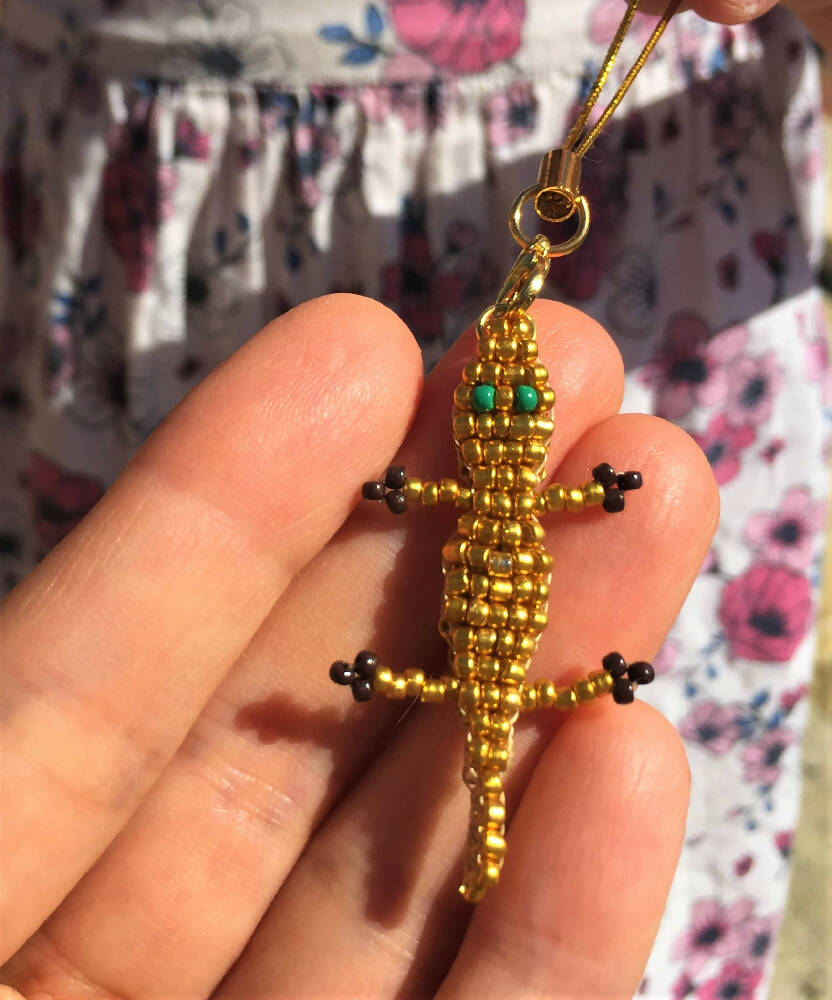 Fingers holding Naryanabeads double layer golden colour crocodile. Body made of shiny golden seed beads, dark green eyes, black paws. Light pink floral dress as background