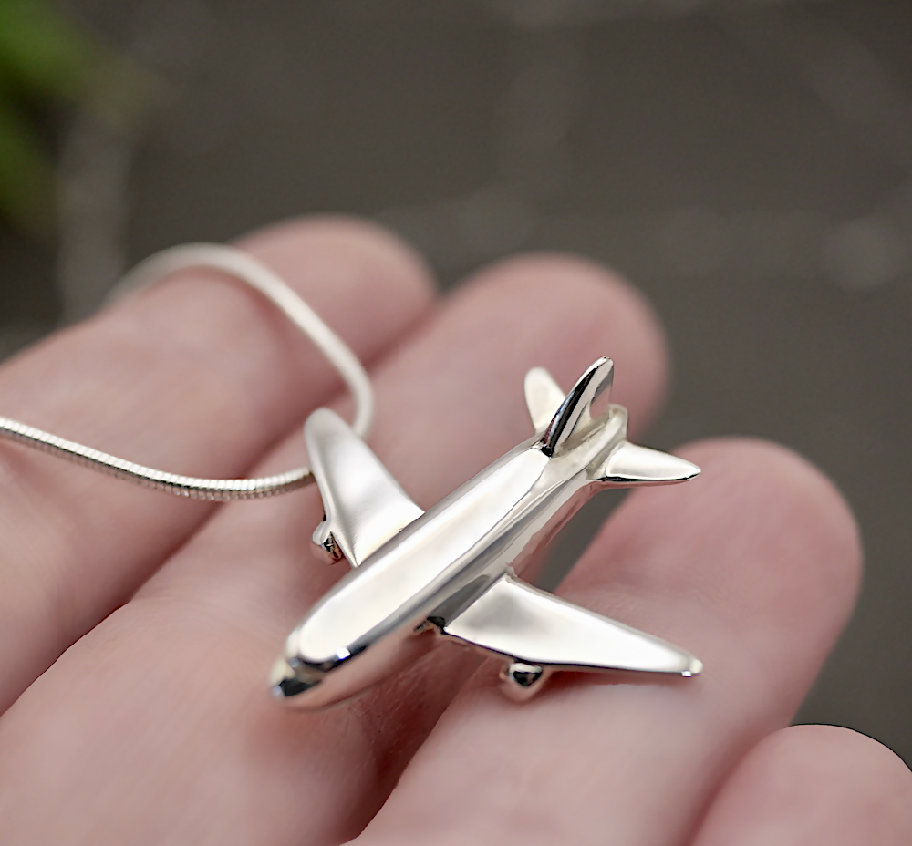 Image of handmade shiny sterling silver aeroplane shaped pendant by Purplefish Designs Jewellery on silver snake chain necklace. The pendant is draped over an open hand to give an indication of size and scale, on a grey marble background